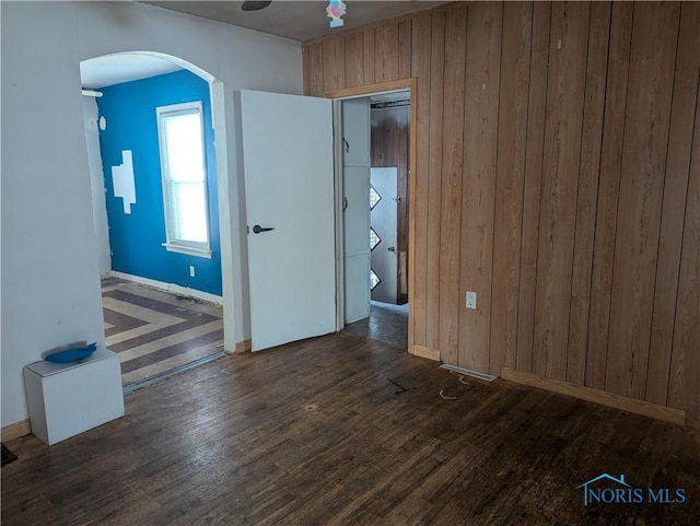 empty room featuring arched walkways, wooden walls, baseboards, and wood finished floors