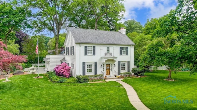 colonial-style house with stucco siding, a chimney, a front lawn, and roof with shingles