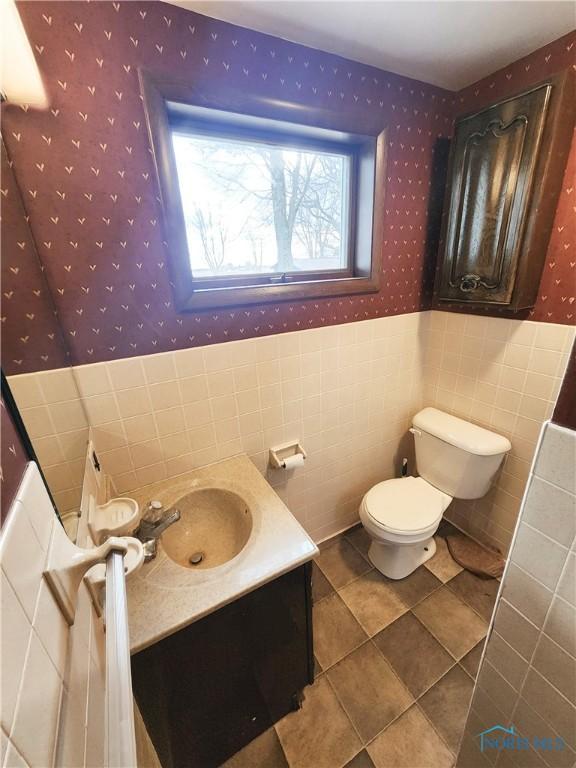 bathroom with wallpapered walls, wainscoting, toilet, vanity, and tile walls