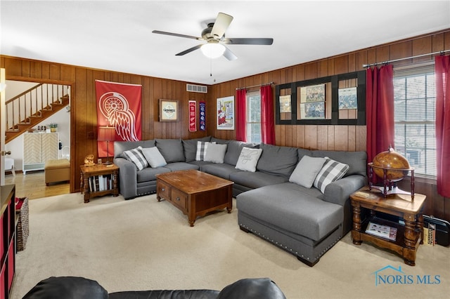 carpeted living area with wooden walls, stairs, visible vents, and a ceiling fan