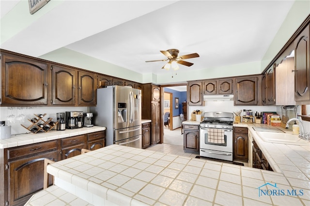 kitchen with dark brown cabinetry, tile counters, stainless steel appliances, under cabinet range hood, and a sink