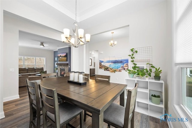 dining room featuring dark wood-type flooring, a stone fireplace, baseboards, and ceiling fan with notable chandelier