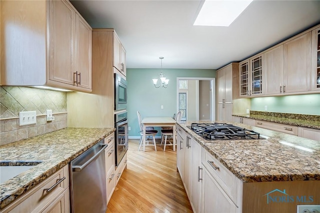 kitchen featuring light wood finished floors, appliances with stainless steel finishes, glass insert cabinets, light stone countertops, and an inviting chandelier