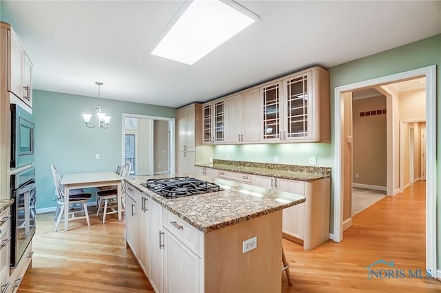 kitchen with a notable chandelier, a kitchen island, appliances with stainless steel finishes, light wood-type flooring, and glass insert cabinets