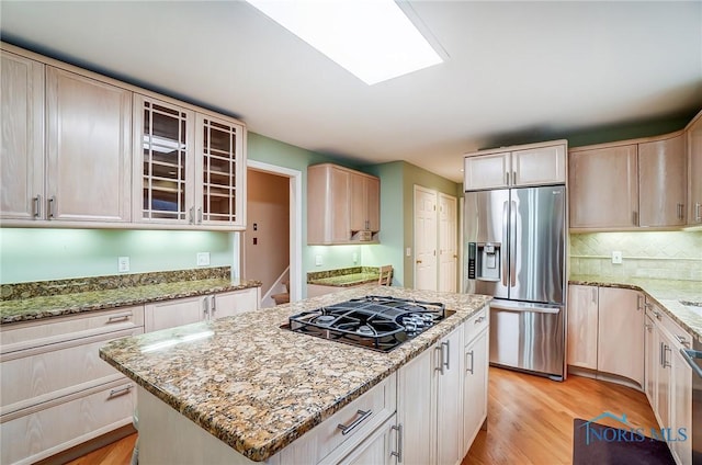 kitchen with appliances with stainless steel finishes, glass insert cabinets, a center island, light stone countertops, and light wood-type flooring