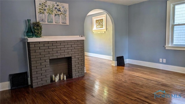 unfurnished living room featuring arched walkways, visible vents, a brick fireplace, baseboards, and hardwood / wood-style flooring