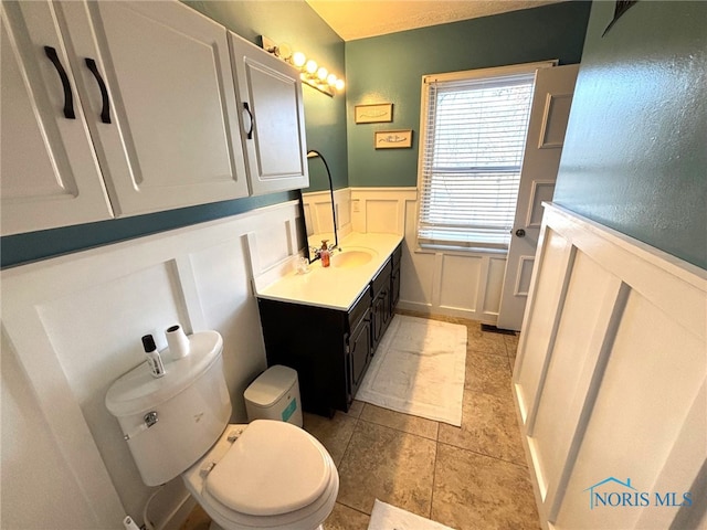 bathroom with a wainscoted wall, toilet, a decorative wall, tile patterned flooring, and vanity