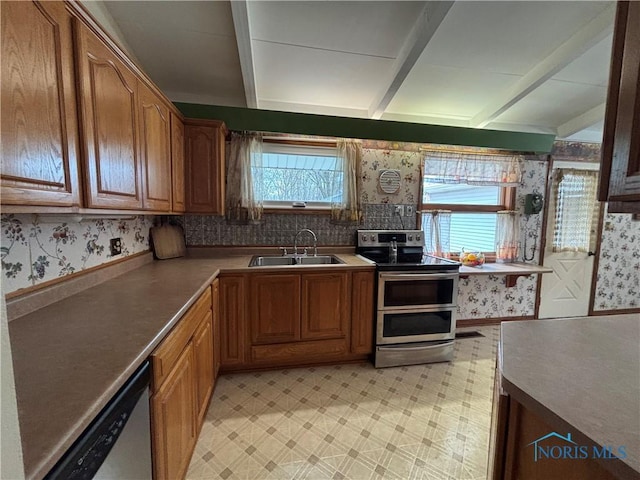 kitchen featuring stainless steel appliances, a sink, light floors, beamed ceiling, and wallpapered walls