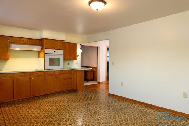 kitchen with under cabinet range hood, baseboards, light countertops, white oven, and brown cabinets