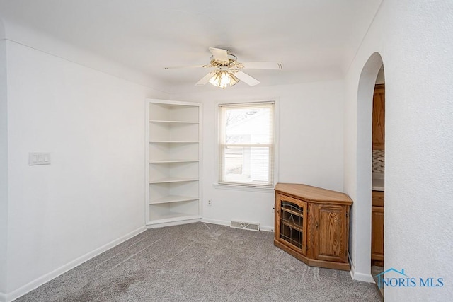 carpeted empty room featuring visible vents, built in shelves, ceiling fan, baseboards, and arched walkways