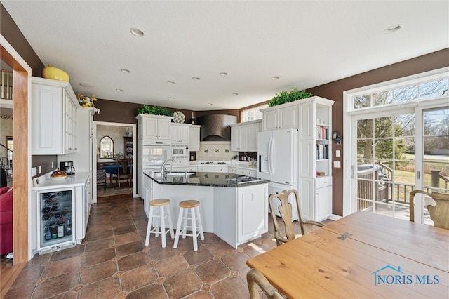 kitchen with an island with sink, a sink, white appliances, wine cooler, and wall chimney range hood