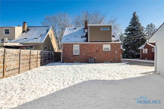 snow covered property featuring brick siding, central AC unit, a chimney, and fence