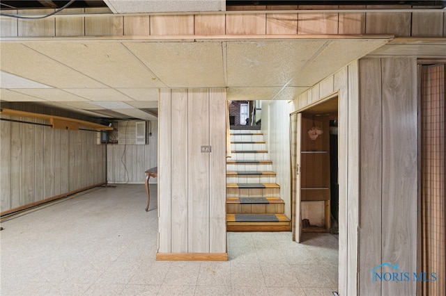 finished basement with tile patterned floors, wooden walls, stairway, and a drop ceiling