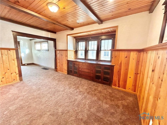 carpeted empty room with wooden walls, a wainscoted wall, visible vents, wooden ceiling, and beamed ceiling