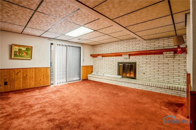 unfurnished living room featuring a wainscoted wall, a drop ceiling, carpet floors, wooden walls, and a brick fireplace