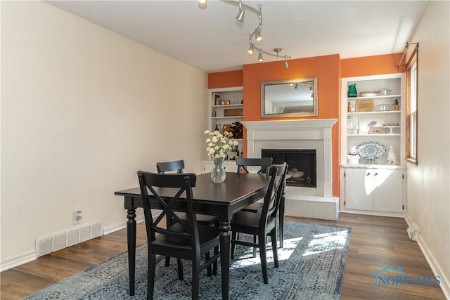 dining room with visible vents, a fireplace with raised hearth, built in features, baseboards, and dark wood-style flooring