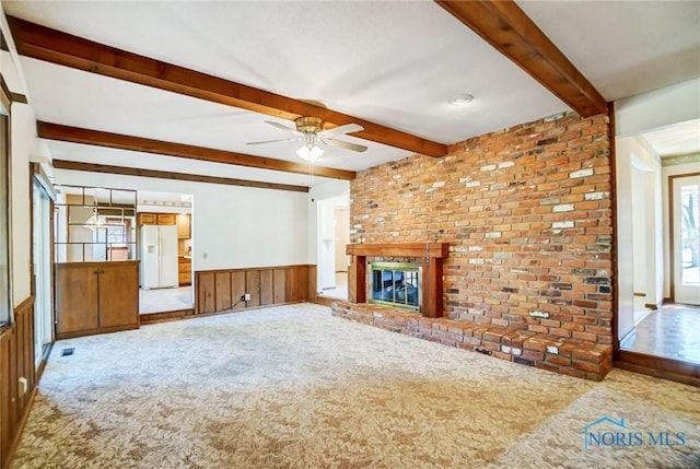 unfurnished living room with beamed ceiling, a wainscoted wall, visible vents, a ceiling fan, and a brick fireplace