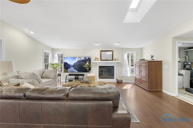 living room featuring a glass covered fireplace, baseboards, vaulted ceiling with skylight, and wood finished floors