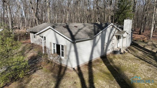 view of side of property with a forest view, central AC, a chimney, and a shingled roof