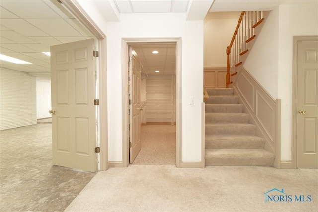 hallway with carpet, a drop ceiling, stairs, wainscoting, and a decorative wall