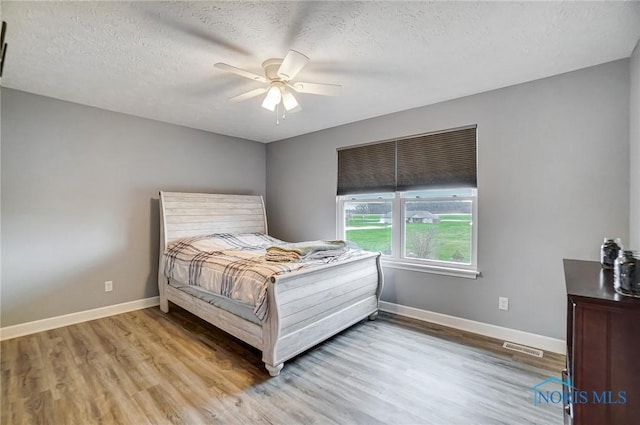 bedroom with a textured ceiling, light wood-type flooring, baseboards, visible vents, and ceiling fan