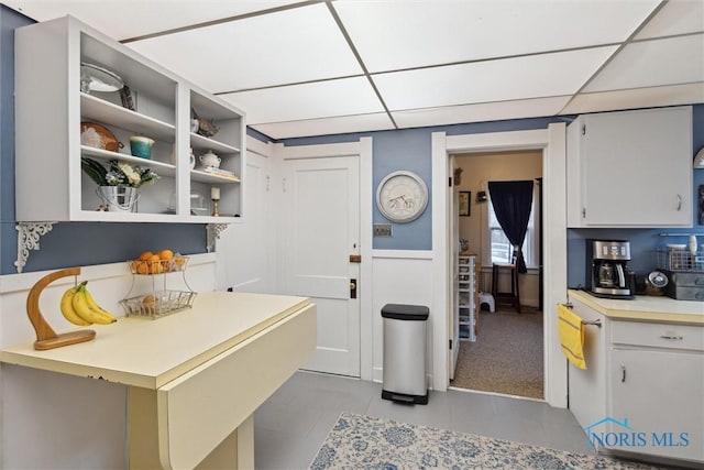 kitchen featuring light tile patterned floors, light countertops, a wainscoted wall, open shelves, and a drop ceiling