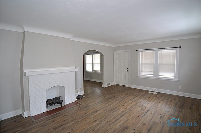 unfurnished living room with arched walkways, visible vents, wood finished floors, a brick fireplace, and baseboards