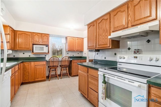 kitchen with backsplash, under cabinet range hood, dark countertops, and white appliances