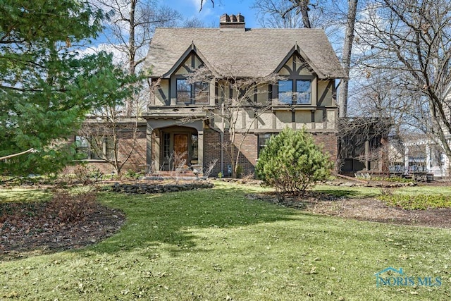 tudor home featuring a shingled roof, a chimney, stucco siding, brick siding, and a front lawn