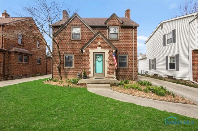 tudor-style house with a front yard, a shingled roof, brick siding, and a chimney