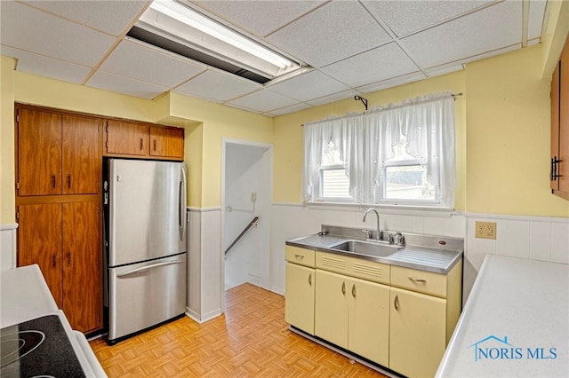 kitchen featuring a paneled ceiling, freestanding refrigerator, stainless steel countertops, a sink, and brown cabinets
