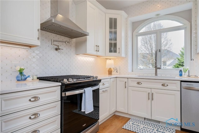kitchen featuring wall chimney exhaust hood, white cabinets, glass insert cabinets, light countertops, and appliances with stainless steel finishes