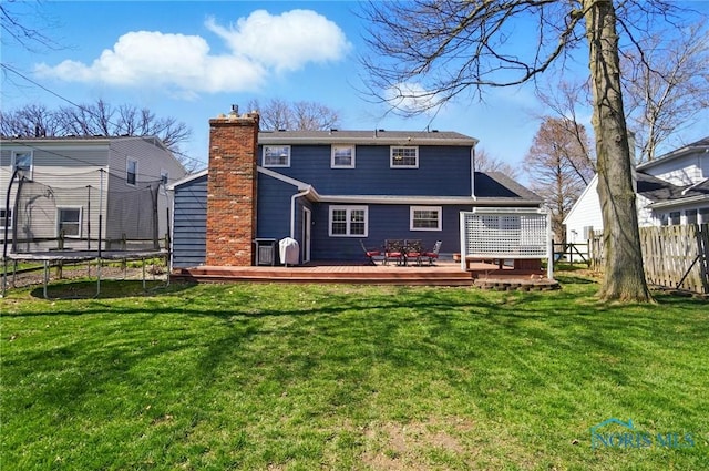 rear view of house with a trampoline, a chimney, fence, a yard, and a wooden deck