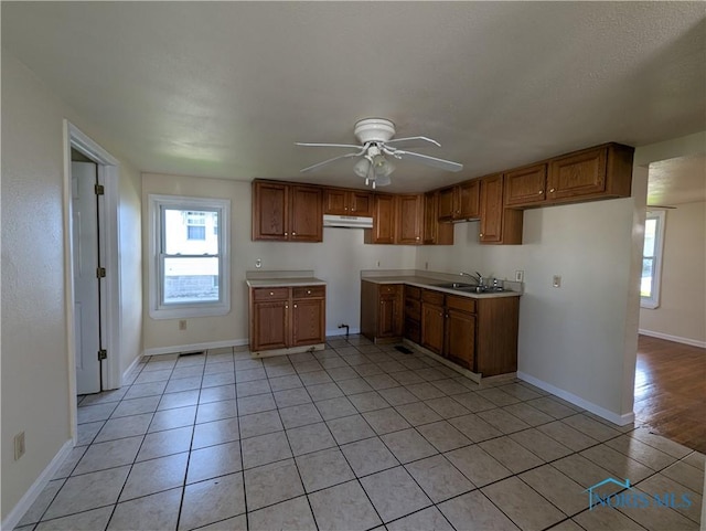 kitchen with a sink, under cabinet range hood, brown cabinetry, a ceiling fan, and baseboards
