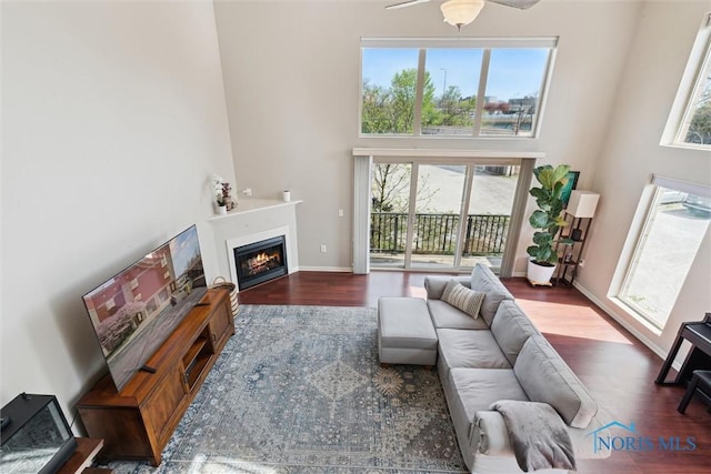 living area featuring a warm lit fireplace, a ceiling fan, baseboards, wood finished floors, and a towering ceiling