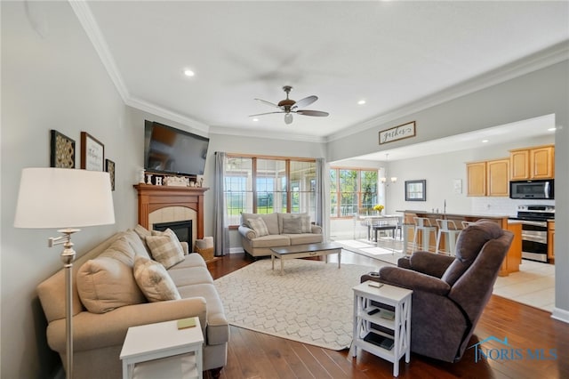 living room with a ceiling fan, crown molding, a tiled fireplace, wood-type flooring, and recessed lighting