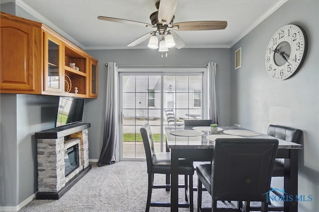 dining space featuring baseboards, carpet, ceiling fan, a stone fireplace, and ornamental molding