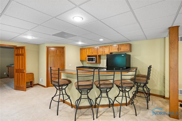 kitchen featuring stainless steel microwave, refrigerator, a kitchen bar, light carpet, and a paneled ceiling