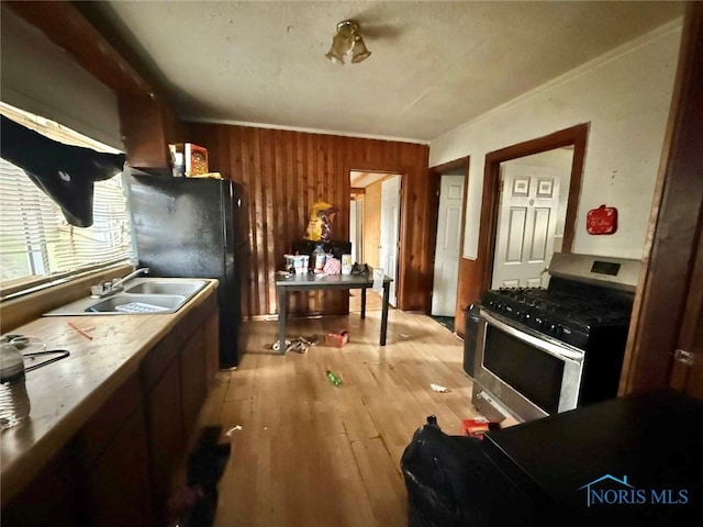 kitchen with a sink, light wood-style floors, freestanding refrigerator, stainless steel gas stove, and wooden walls