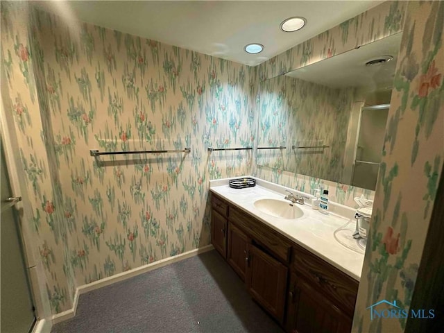 bathroom featuring a sink, a shower with shower door, double vanity, baseboards, and wallpapered walls