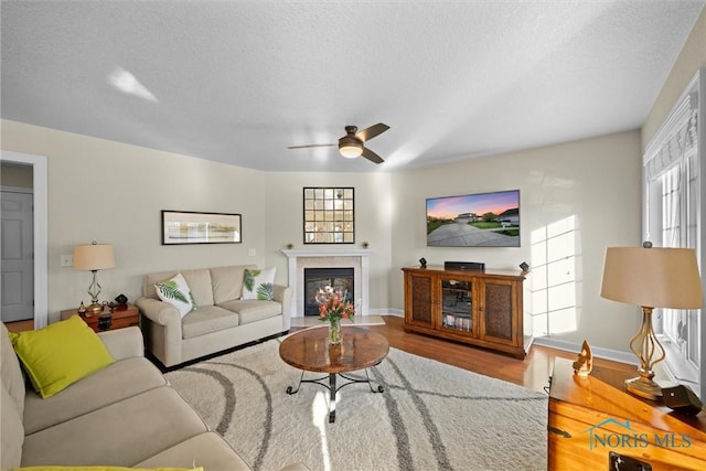 living room featuring ceiling fan, a textured ceiling, wood finished floors, baseboards, and a glass covered fireplace