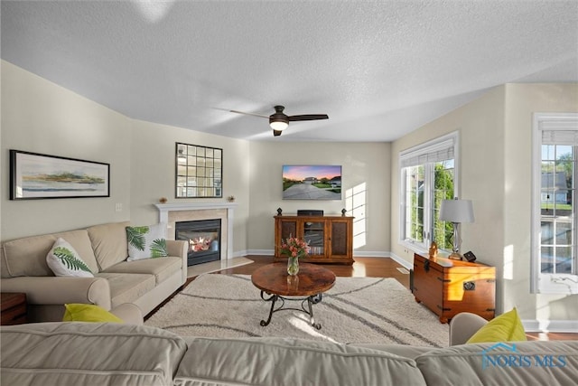 living area with ceiling fan, a textured ceiling, a fireplace with flush hearth, wood finished floors, and baseboards