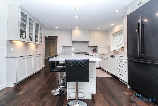 kitchen with glass insert cabinets, a kitchen bar, high end black refrigerator, dark wood finished floors, and light countertops