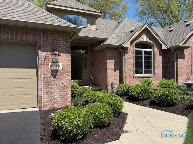 view of exterior entry with a garage, a shingled roof, and brick siding