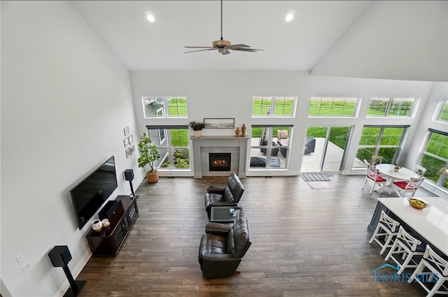 living area with high vaulted ceiling, healthy amount of natural light, ceiling fan, a lit fireplace, and dark wood-style flooring