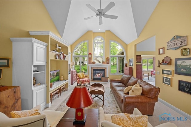 living area featuring a ceiling fan, baseboards, a glass covered fireplace, and high vaulted ceiling