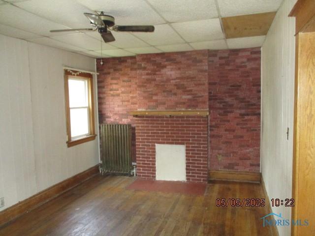 empty room featuring wood finished floors, a ceiling fan, a fireplace, a paneled ceiling, and radiator heating unit