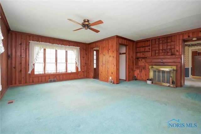 unfurnished living room with carpet, a glass covered fireplace, ceiling fan, visible vents, and wood walls