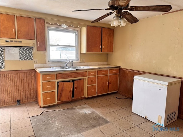 kitchen with white refrigerator, under cabinet range hood, a sink, wainscoting, and brown cabinetry