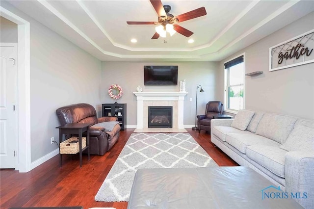 living area with ceiling fan, a raised ceiling, dark wood-type flooring, a tiled fireplace, and baseboards